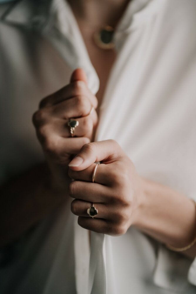 Close-up of hands adjusting a white shirt, wearing rings with a soft, blurred background.
