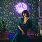Woman in a teal dress sits on a purple sofa in a plant-filled room, with a glowing purple aura above her head suggesting magic or energy.