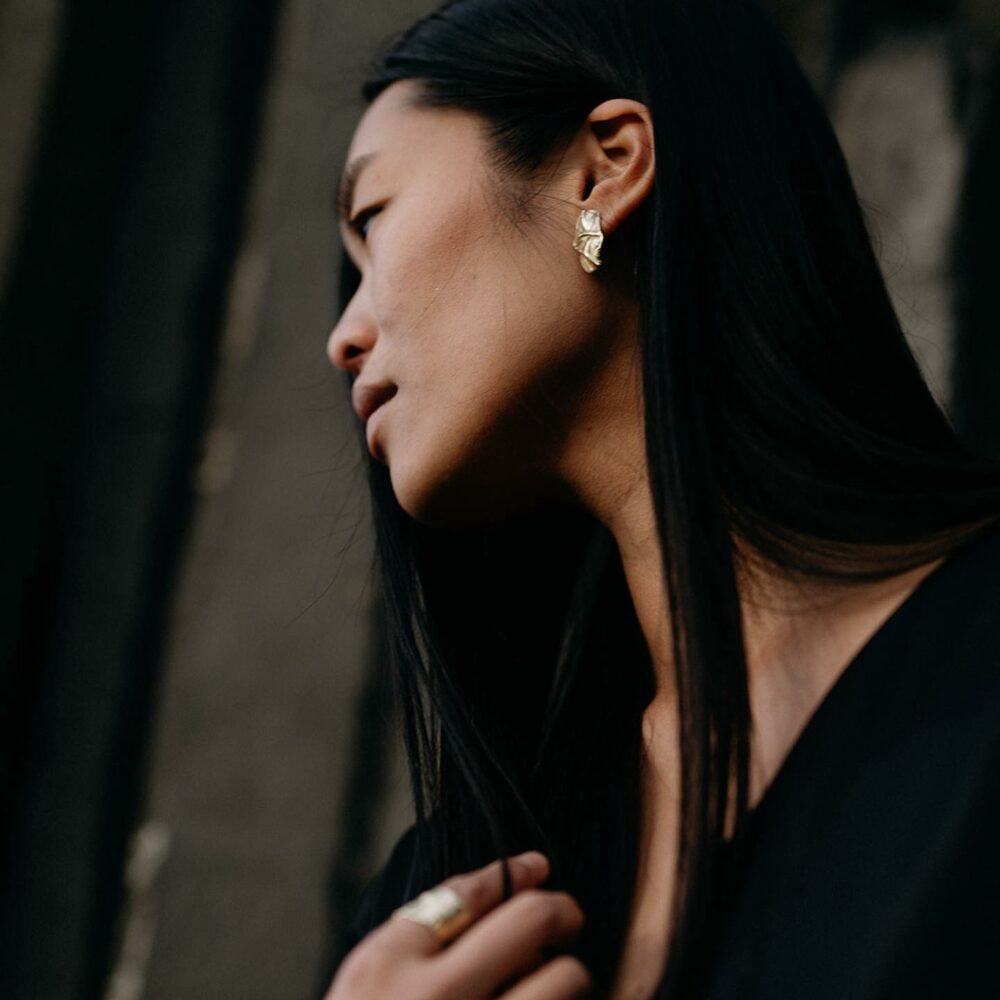 Close-up side profile of a woman with long black hair wearing a gold leaf-shaped earring in a dark setting.