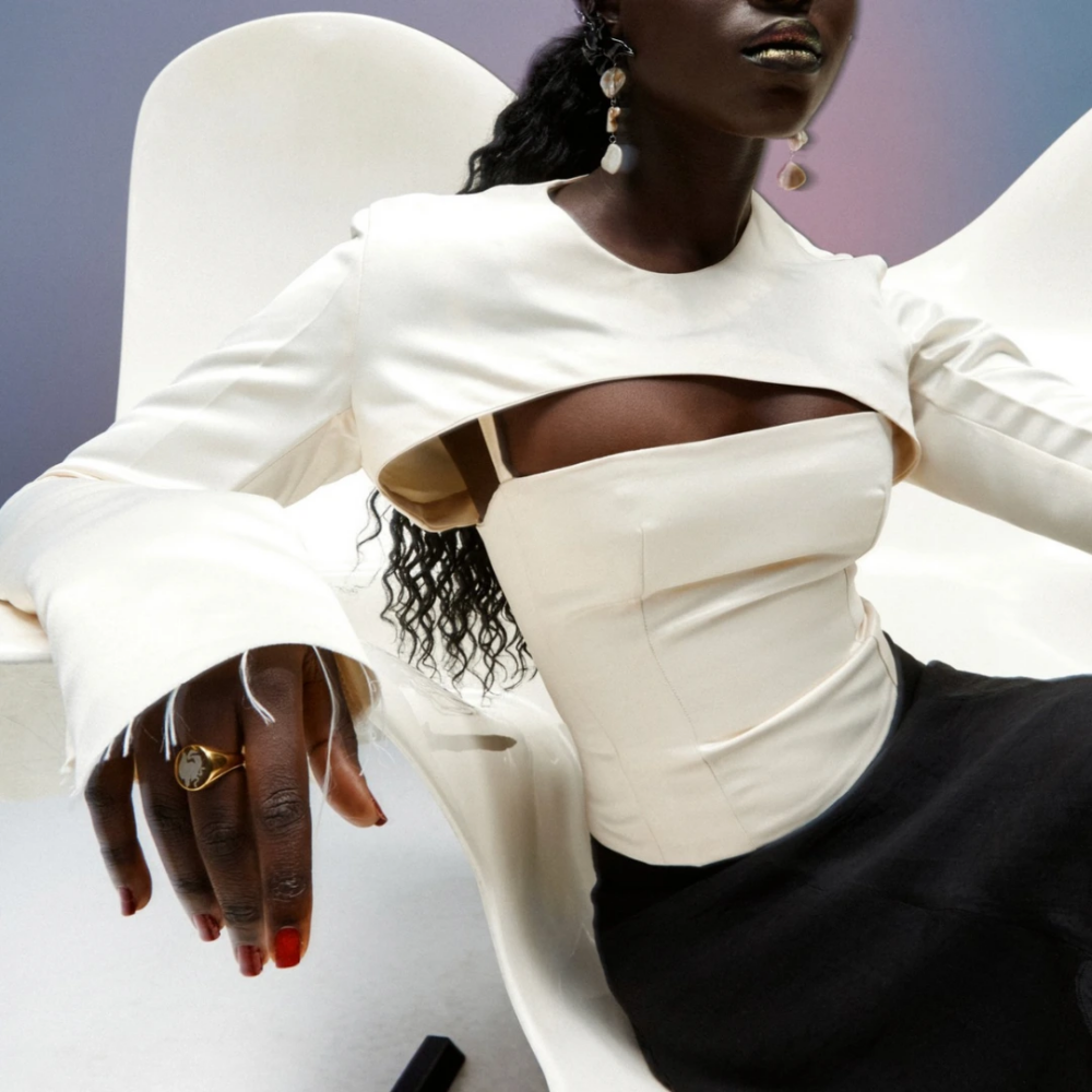 Fashion editoral shot of a dark-skinned model in a white sculptural top with a cut-out neckline, sitting on a white chair, accessorized with dangling earrings and a gold ring on her finger.