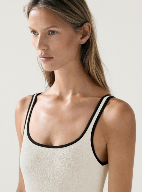 Woman with light brown hair wearing a beige tank top with black trim, looking to the left.