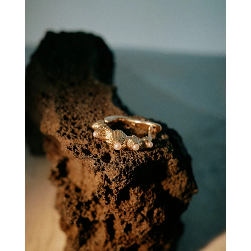 Gold bracelet with small pearls rests on a dark rocky surface at the beach by the sea edge.