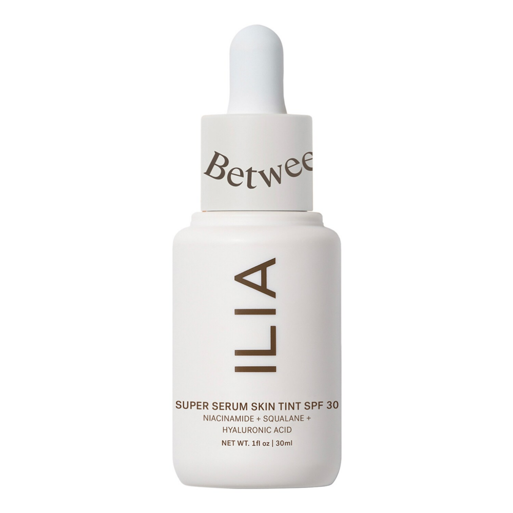 Close-up of a white skincare dropper bottle (ILIA) with a rounded dropper cap and brown branding text visible on the label, against a white background.
