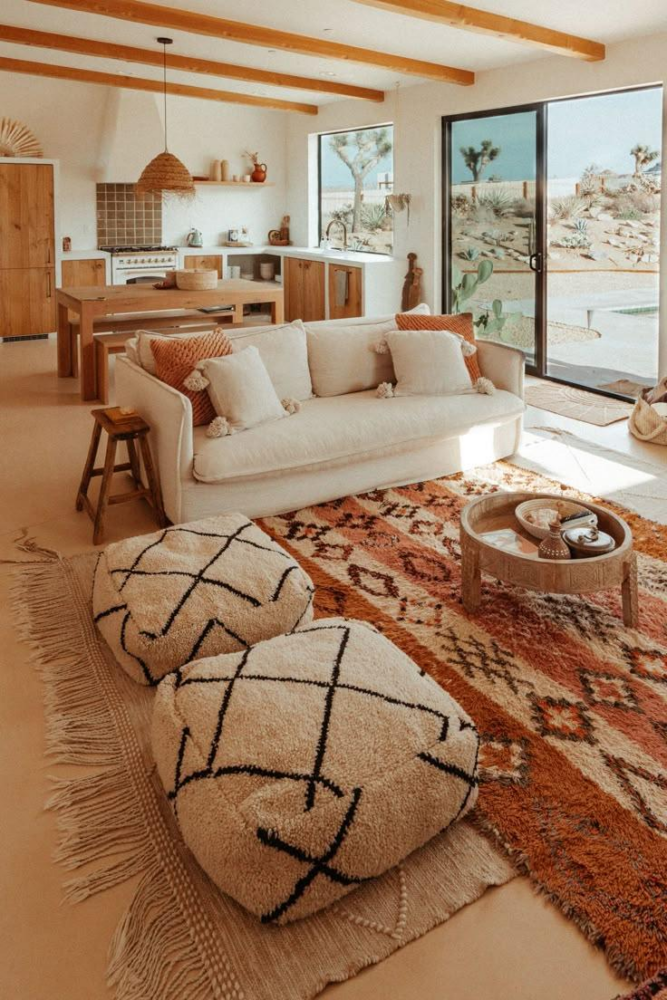 Open-plan living room with a white sofa, patterned rug, and woven poufs; sliding glass doors reveal a desert garden outside the house.