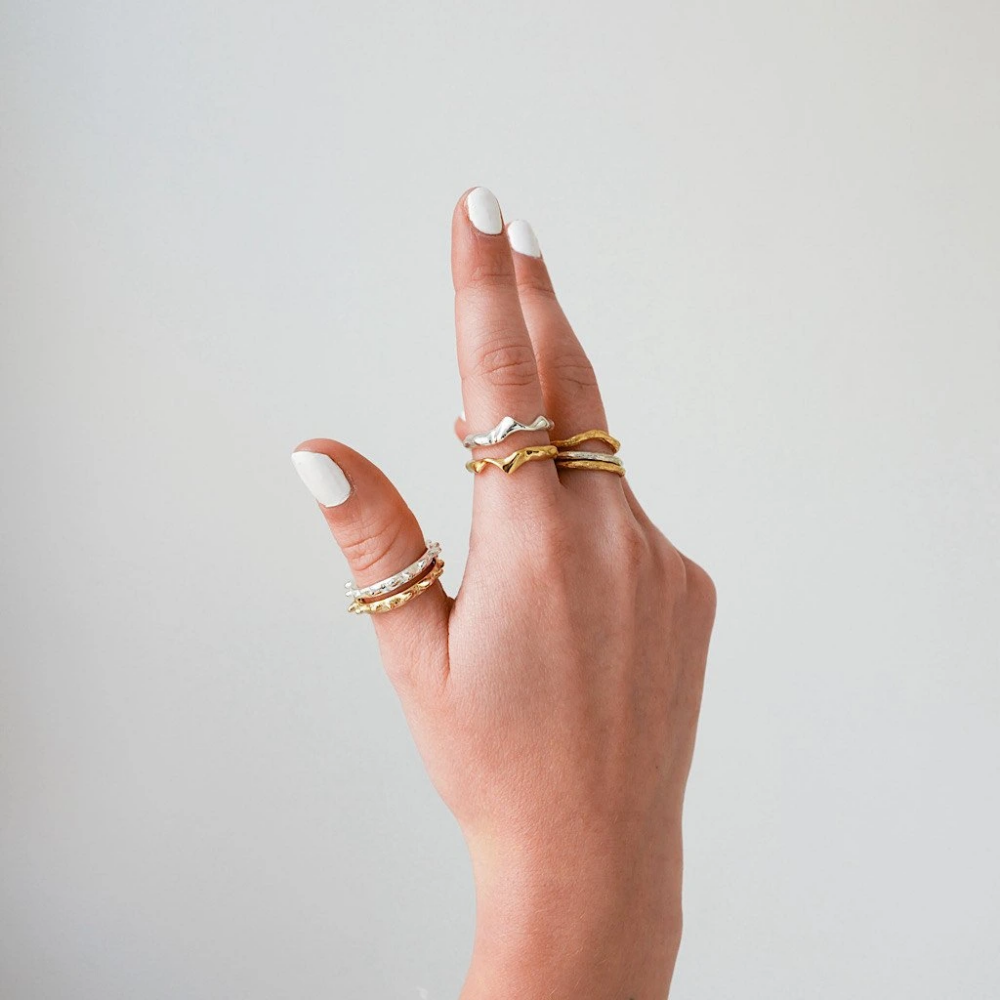 Close-up of a hand with white nail polish wearing several gold and silver stacked rings against a pale background.
