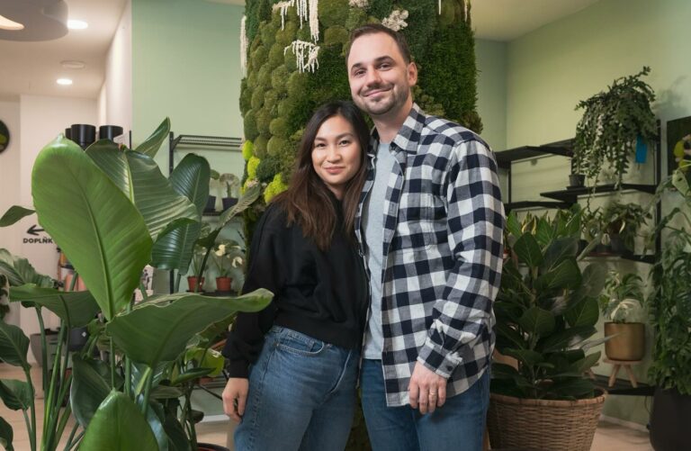 Smiling couple posing in a plant shop, surrounded by large potted plants and a moss-covered wall behind them.