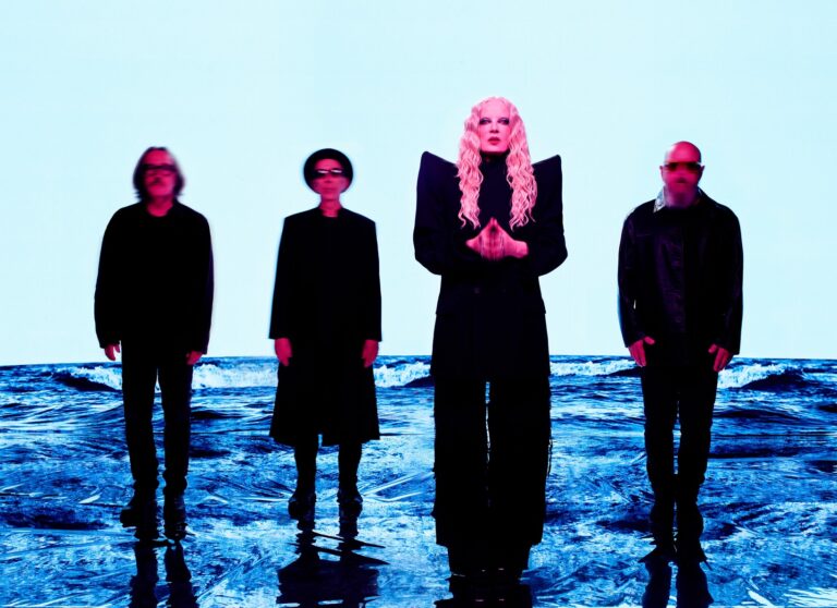 Group of five models standing in shallow water against a pale blue backdrop; central figure with long pink hair wearing a black outfit, flanked by others in dark clothing.