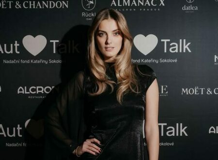 Woman in a sleek black dress posing on a dark backdrop featuring sponsor logos at an event.