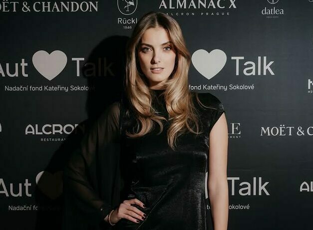 Woman in a sleek black dress posing on a dark backdrop featuring sponsor logos at an event.