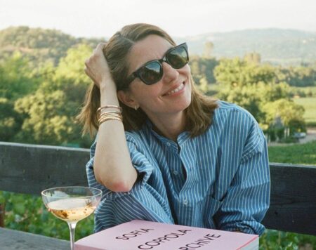 Smiling woman in sunglasses sits at a wooden outdoor table with a pink 'Sofia Coppola Archive' book and a wine glass nearby.