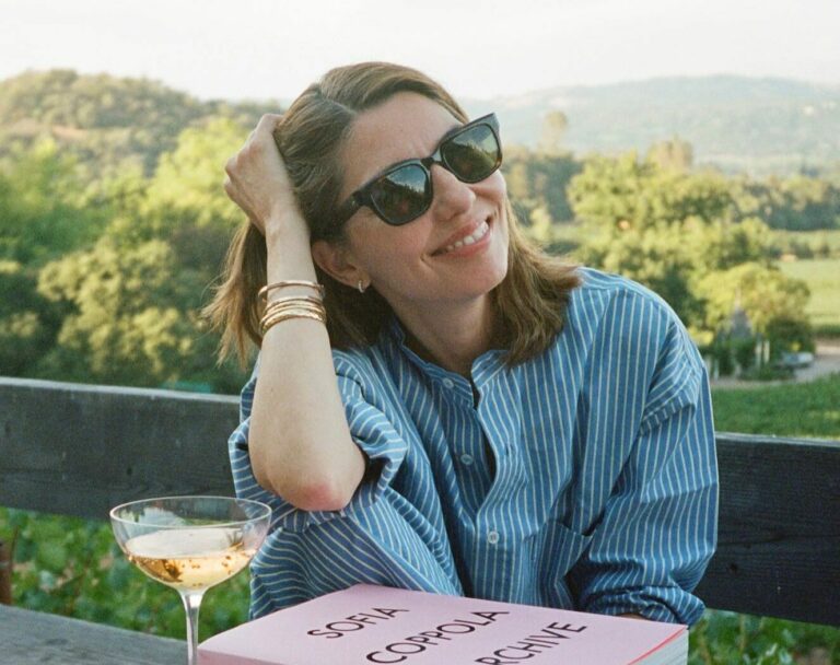 Smiling woman in sunglasses sits at a wooden outdoor table with a pink 'Sofia Coppola Archive' book and a wine glass nearby.