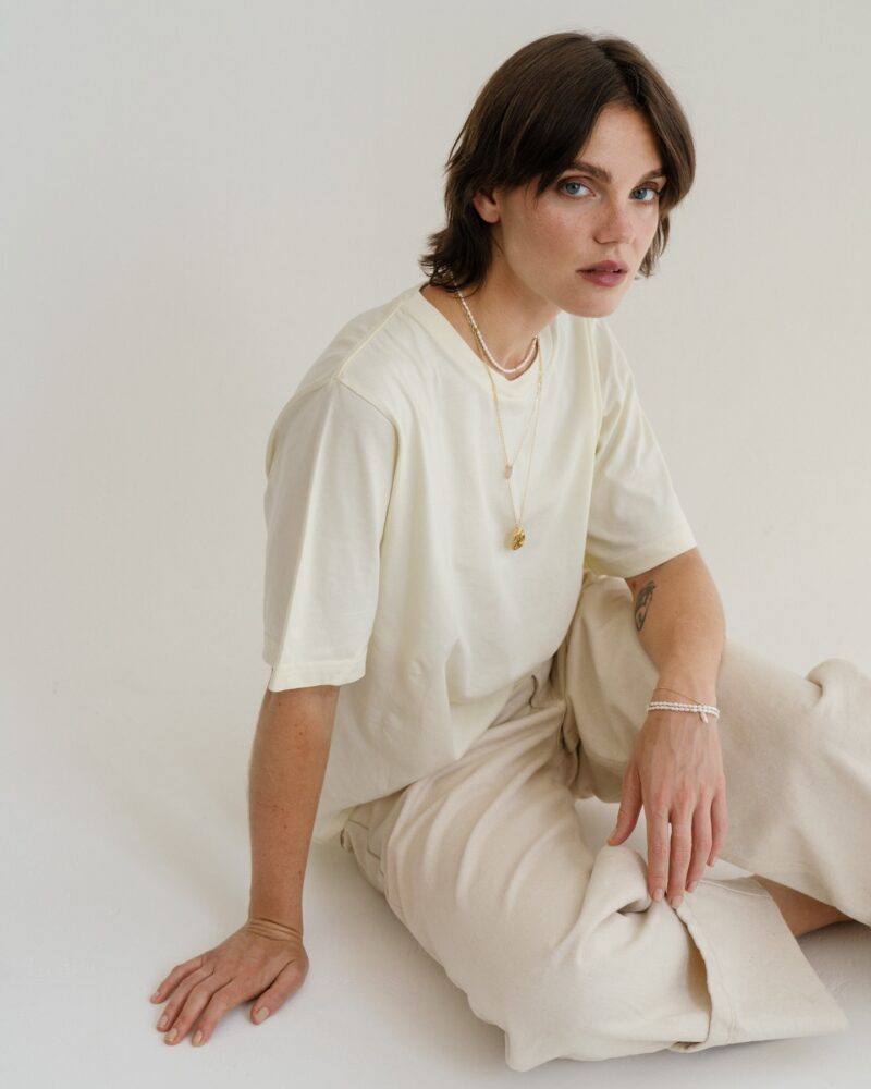 Person with short brown hair wearing a cream T-shirt and beige pants, seated on the floor against a pale wall, accessorized with layered necklaces and bracelets.