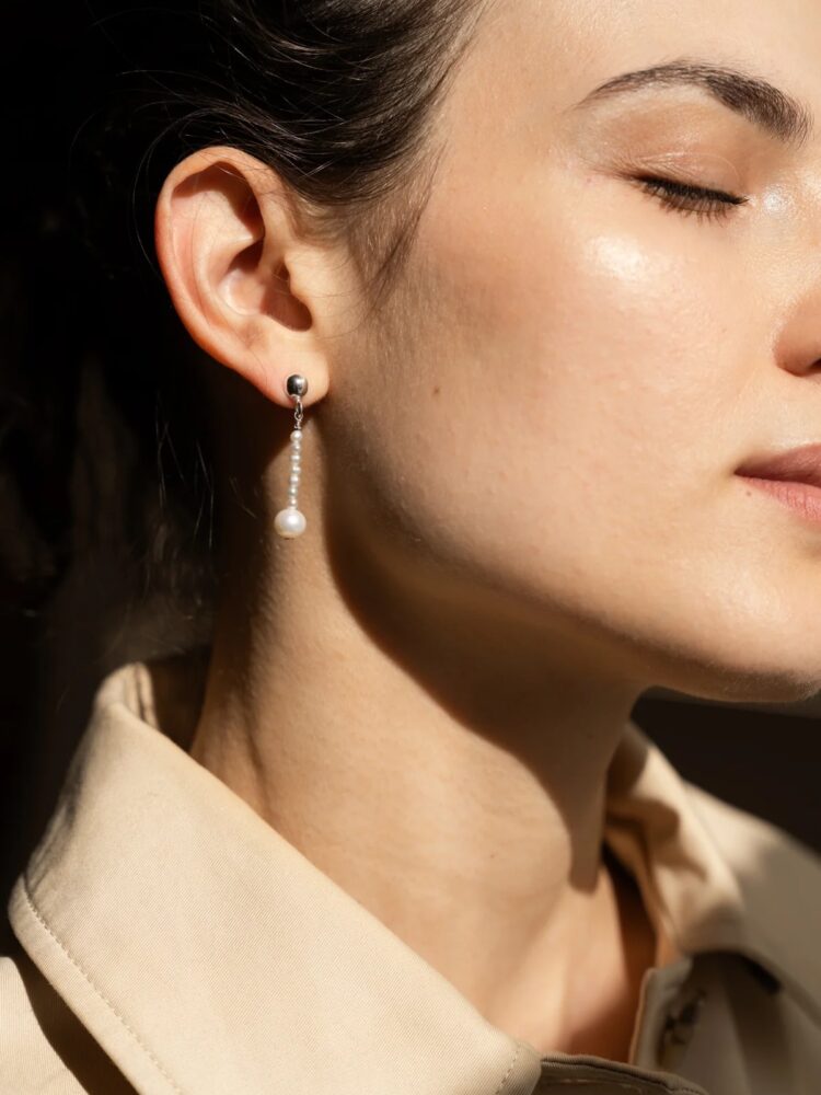 Close-up of a woman's left ear wearing a delicate pearl drop earring with small beads and a silver stud, against a beige collared shirt.