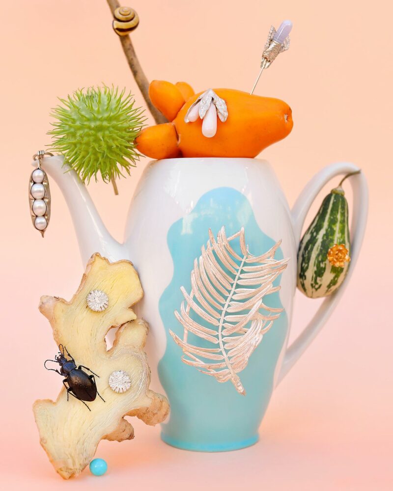 Whimsical still-life collage: a turquoise teapot with gold fern pattern, topped by an orange carrot, green spiky seed pod, beetle on wood slice, and turquoise bead on a peach background.