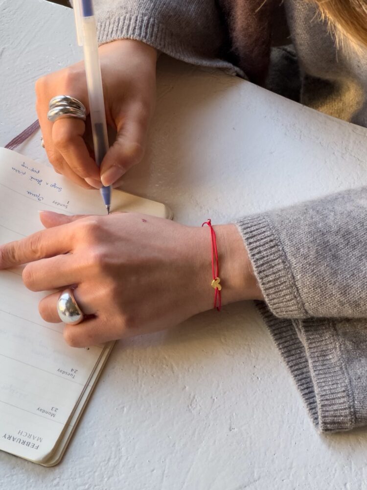 Close-up of hands writing in a notebook with a blue-ink pen; wearing silver rings and a red string bracelet.