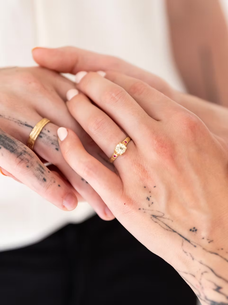 Close-up of two hands interlaced, showing a gold engagement ring with a diamond on one finger and a plain gold band on the other, with a tattoo visible on the forearm.