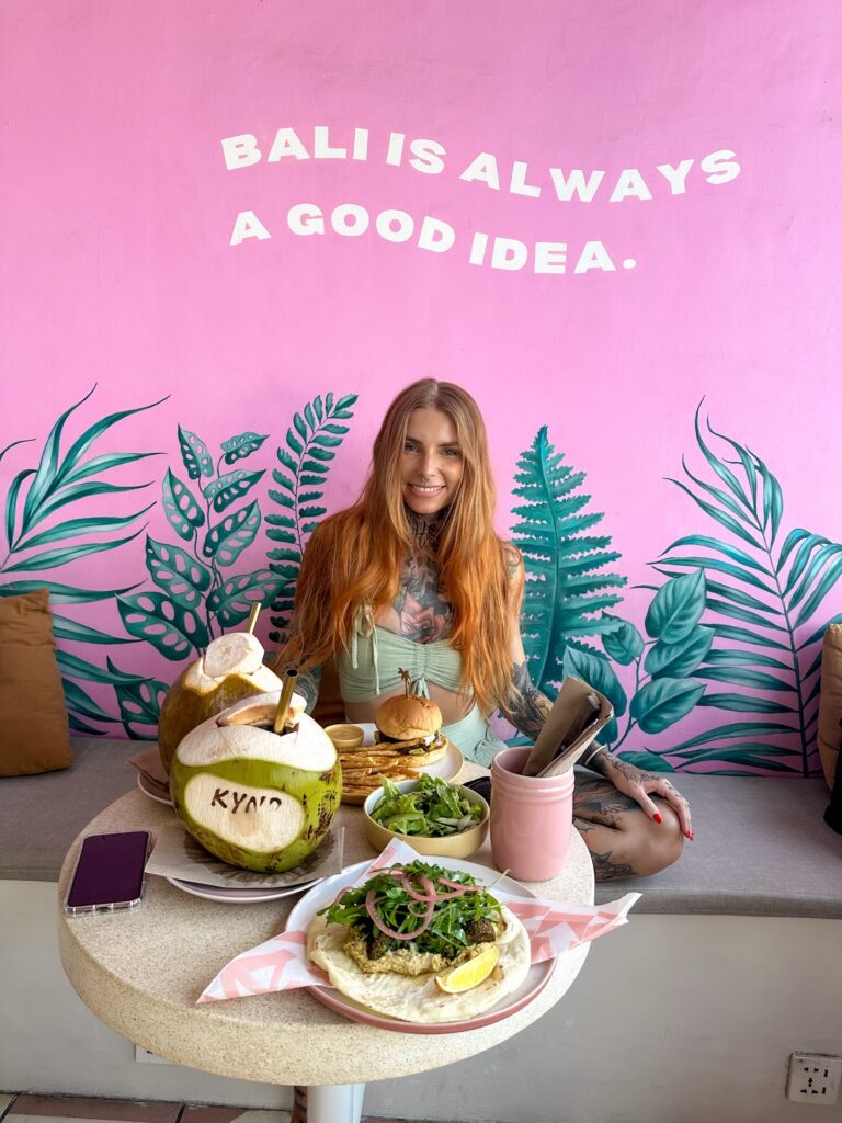 Woman with long hair sitting at a pink mural reading 'BALI IS ALWAYS A GOOD IDEA,' with tropical leaf artwork and coconut drinks on the table