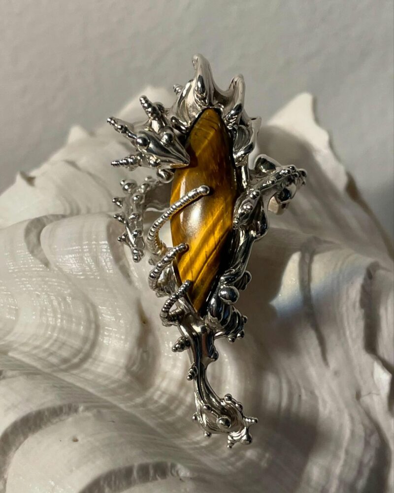 Close-up of a silver ornate ring featuring a yellow-brown tiger's eye gemstone, surrounded by intricate sculpted metal branches, resting on white tissue paper.