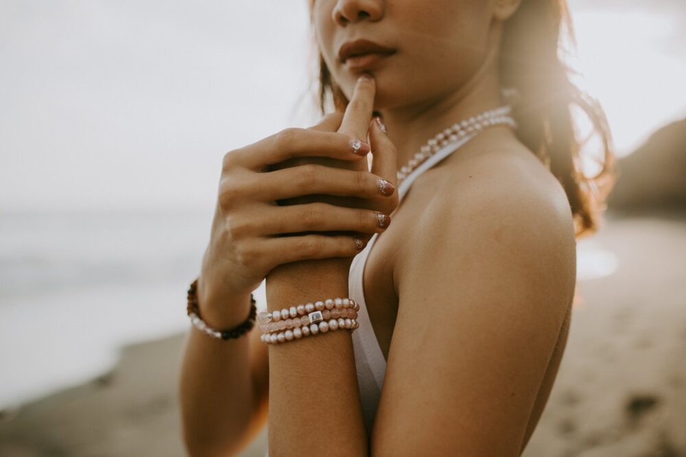 Close-up of a woman at the beach wearing a white swimsuit and multiple pearl bracelets, with her fingers resting near her lips.
