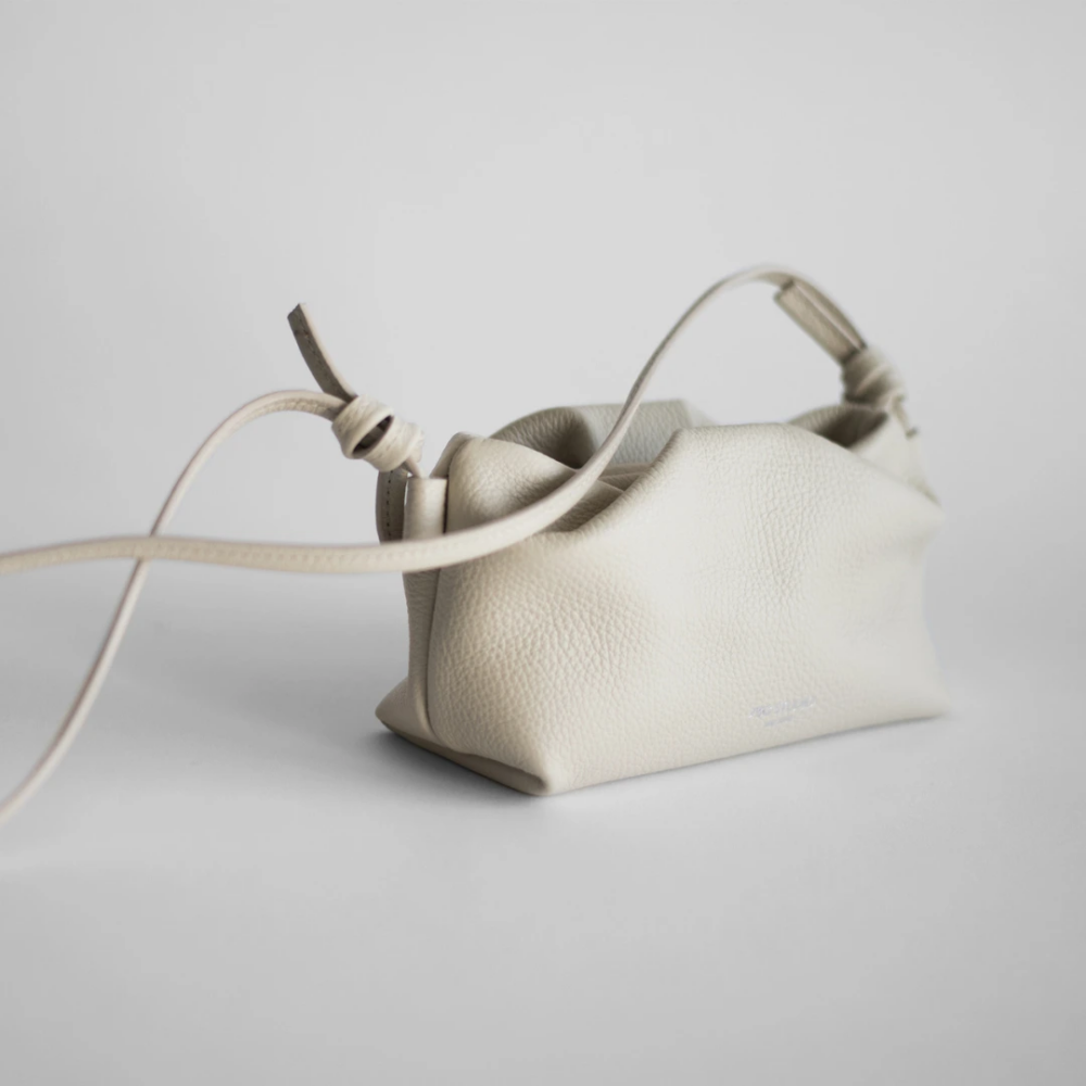 Cream leather crossbody bag with a long knotted strap on a pale backdrop, close-up details visible on textured surface.