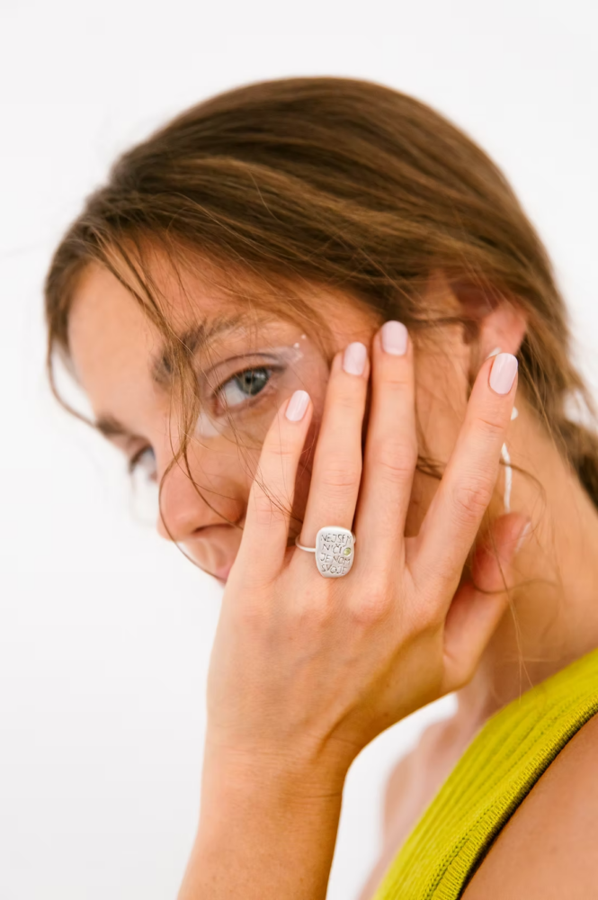 Close-up of a woman with light brown hair covering her face with her left hand, showing a silver signet ring and pale pink nails, wearing a lime-green top.