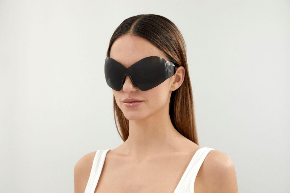 Woman wearing oversized black protective goggles or sleep-mask-style eyewear, facing slightly left in a white sleeveless top