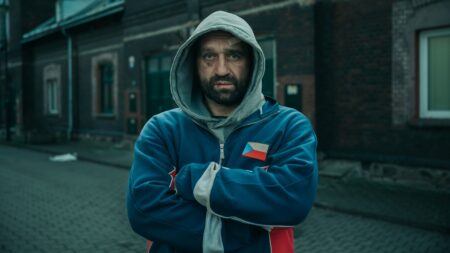 Bearded man in a blue hoodie with arms crossed on a cobblestone street, urban bricks behind him, a bandage on his forehead edge visible.
