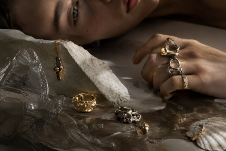 Close-up of a hand with chunky rings resting on a stone slab, surrounded by gold and silver jewelry and a seashell, in moody lighting.