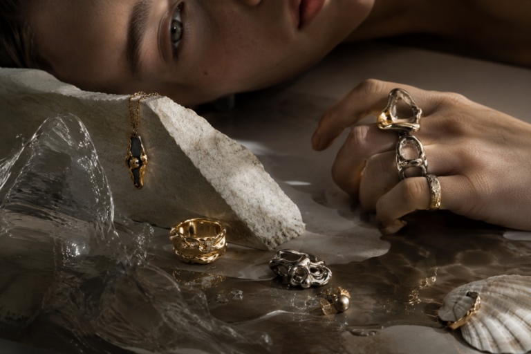 Close-up of a hand with chunky rings resting on a stone slab, surrounded by gold and silver jewelry and a seashell, in moody lighting.