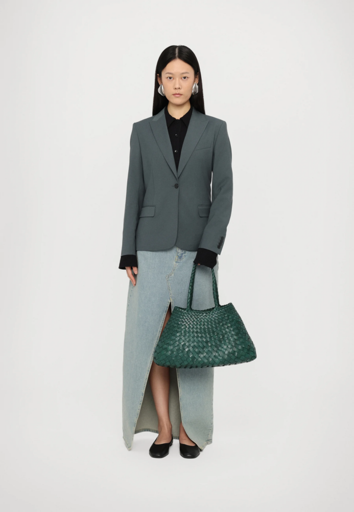 Woman in a gray blazer over a black blouse, wearing a long light-wash denim skirt with a front slit, black flats, and carrying a green woven tote against a white backdrop.