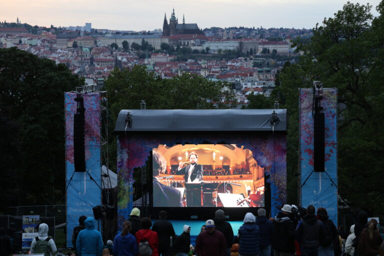 Outdoor concert stage with a large screen showing a performer, banners on the sides, and a distant city skyline framed by trees on both sides.