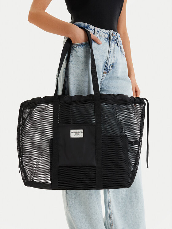 Black mesh tote bag with solid front pocket and two long shoulder straps being held by a person in light-wash jeans and a black top; label reads 'GUESS JEANS' on the front pocket
