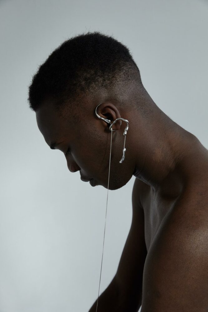 Close-up side profile of a person with short curly hair, wearing a silver ear cuff and dangling chain.
