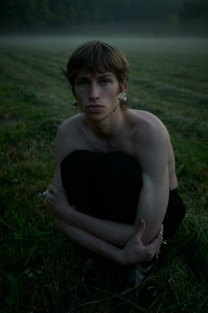Young person sitting shirtless in a grassy field at dusk, arms wrapped around knees, wearing a nose ring and earrings.