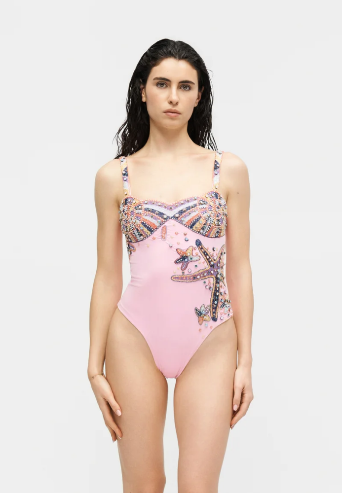 Woman wearing a pink embellished one-piece swimsuit with beadwork and starfish design, standing against a light background.