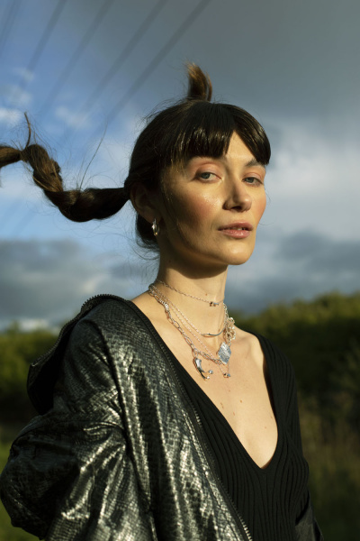 Portrait of a woman outdoors in a black V-neck top and layered necklaces, wind blowing her hair with a metallic jacket over one shoulder.