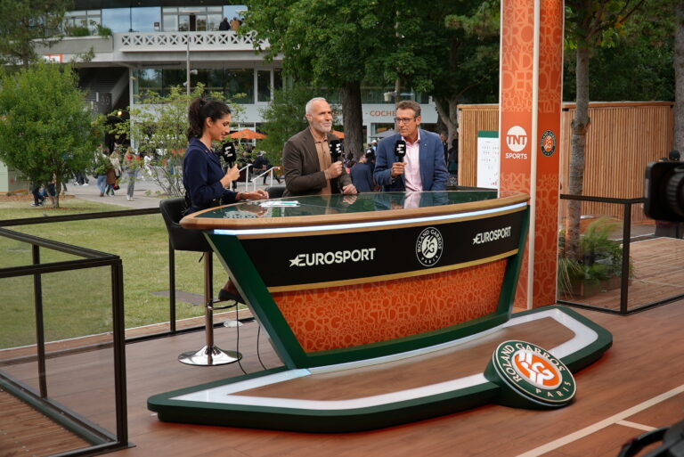 Three TV presenters sit behind a curved broadcast desk with Eurosport logos, holding microphones during an outdoor event with orange banners and trees in the background.