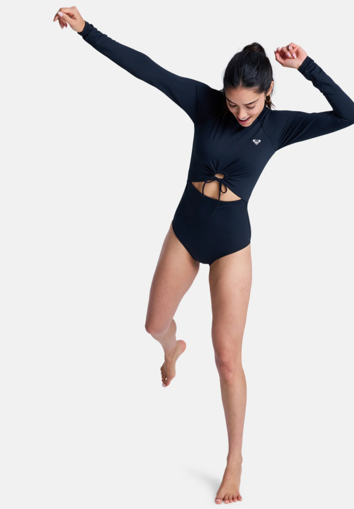 Woman in a navy wetsuit-style swimsuit jumping mid‑air with arms raised, white background, playful pose.
