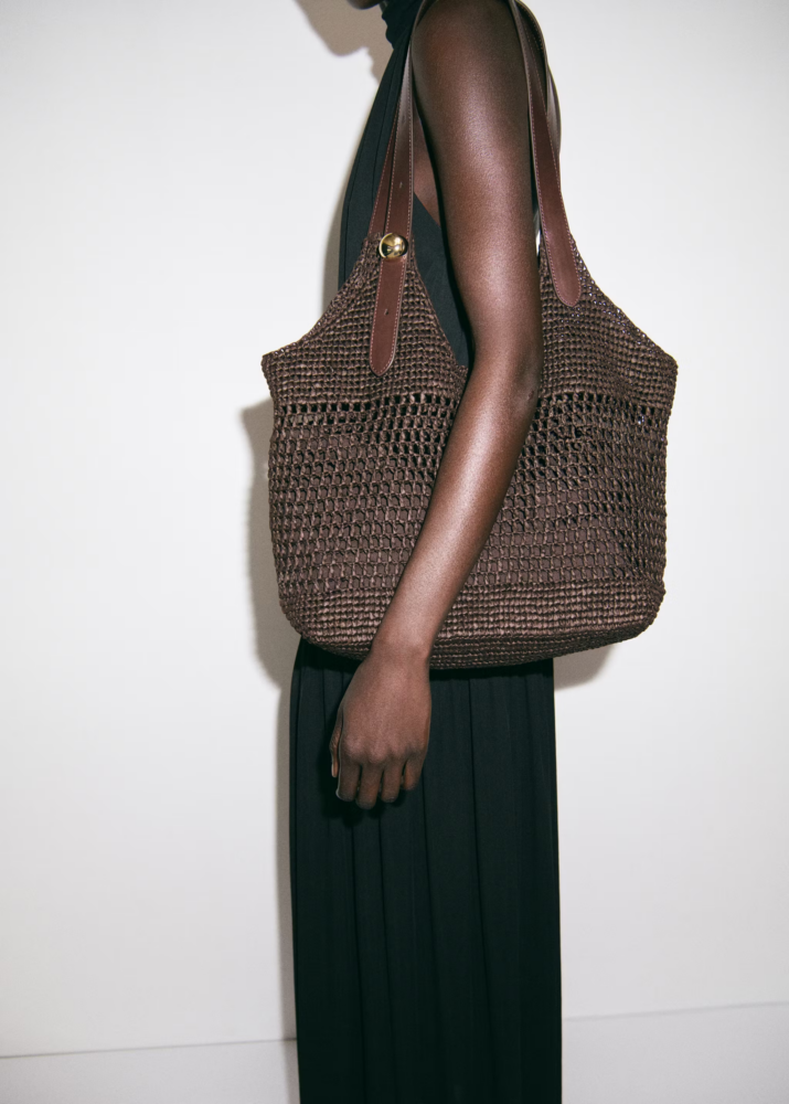 Side view of a person carrying a large brown woven tote bag with leather straps against a black dress and light background, emphasizing the bag.