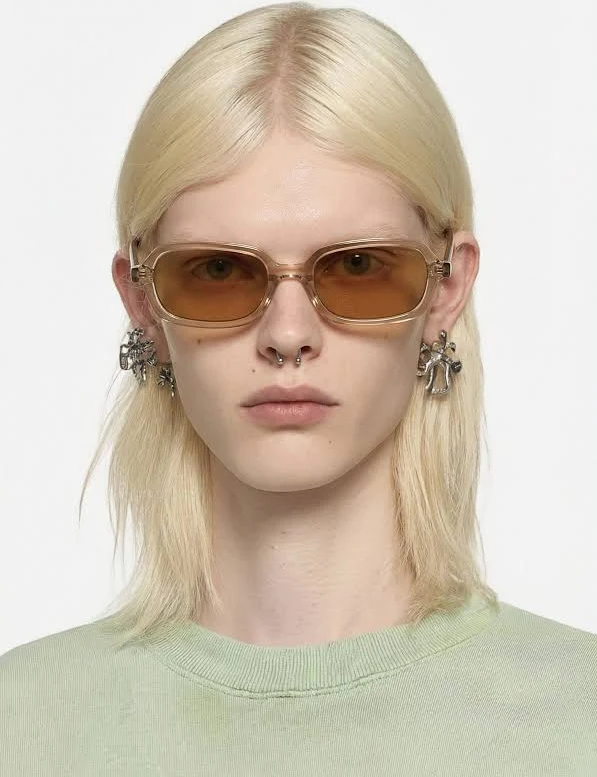 Person with platinum blonde hair wearing brown-tinted sunglasses, septum piercing, and statement earrings against a pale backdrop.