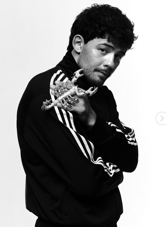 Black-and-white portrait of a man in a striped tracksuit holding an ornate curved dagger across his chest, looking at the camera.