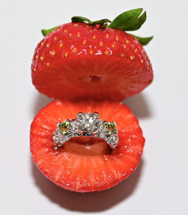 Diamond engagement ring with green stones set in a silver band placed between two halves of a sliced strawberry.