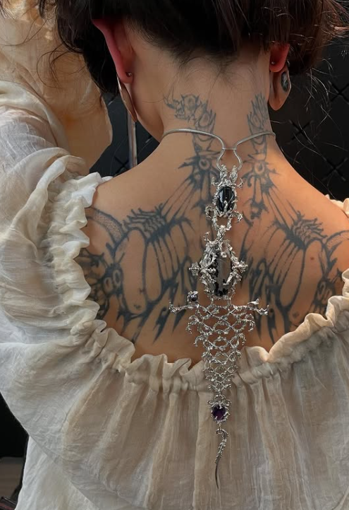 Back view of a person with a large wing tattoo across the shoulders and spine, wearing an ornate silver necklace with a purple gemstone.