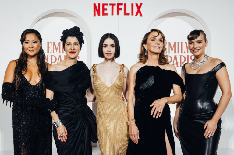Five women in formal gowns pose together on a red carpet with Netflix branding in the background, at an Emmy event scatter.