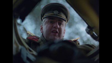 Close-up of an older man in a military uniform looking down at the camera from a cockpit or vehicle interior.