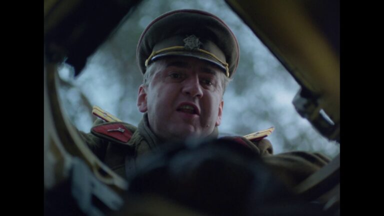 Close-up of an older man in a military uniform looking down at the camera from a cockpit or vehicle interior.