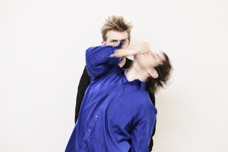 Man in a blue shirt recoils as another man behind him reaches toward his face; motion blur suggests impact in a studio setting.
