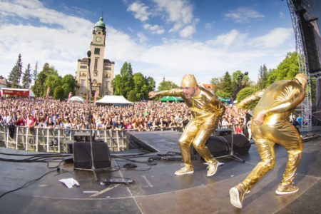 Large outdoor concert crowd watches gold-clad performers on stage under a blue sky.