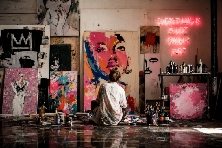 Artist sits on the floor of a paint-splattered studio, working on a large colorful portrait among other artworks.