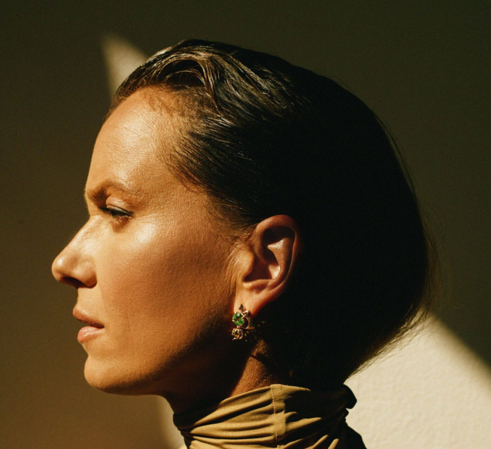 Side profile of a woman with dark hair wearing green gemstone earrings and a brown scarf.